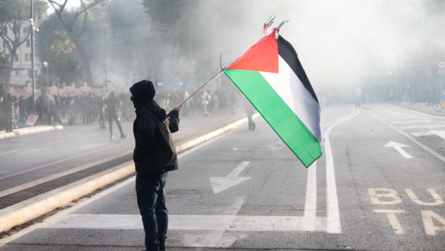 A demonstrator, alone, waves the Palestinian flag, while smoke from gas tanks drift around him